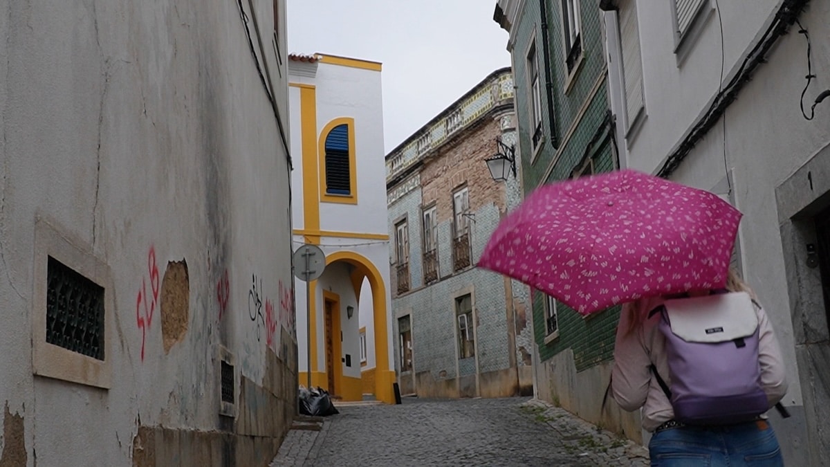 Eine Person mit einem rosafarbenen Regenschirm und einem Rucksack geht an einem bewölkten Tag eine schmale Kopfsteinpflasterstraße entlang, die von alten Gebäuden gesäumt ist.