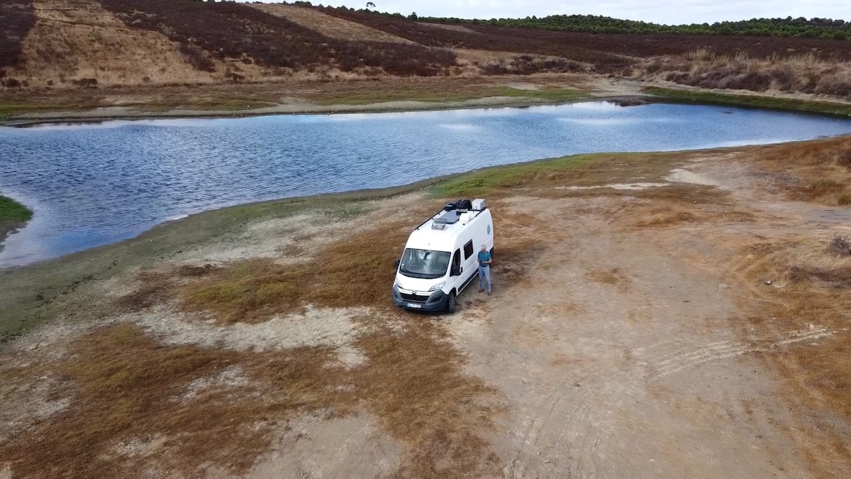 Ein weißes Wohnmobil ist auf trockenem, grasbewachsenem Boden in der Nähe eines Gewässers geparkt, daneben steht eine Person. Im Hintergrund sind Hügel und spärliche Vegetation zu sehen.