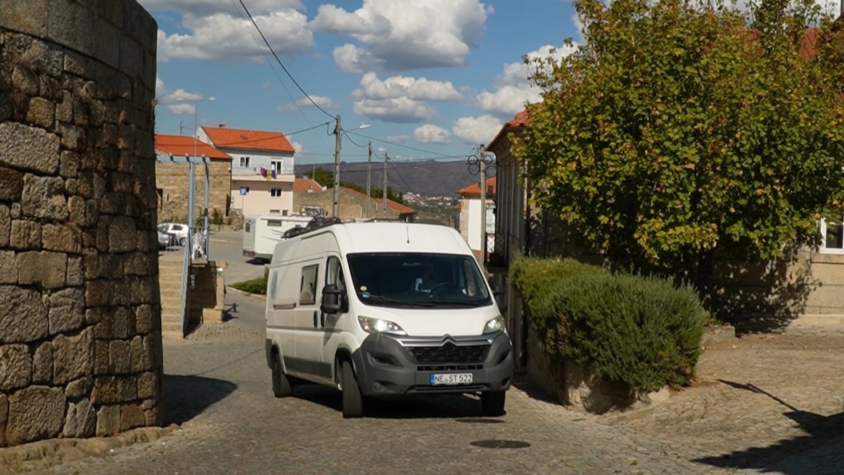 Ein weißer Van fährt auf einer schmalen, von Steinmauern und Gebäuden gesäumten Kopfsteinpflasterstraße unter einem teilweise bewölkten Himmel.
