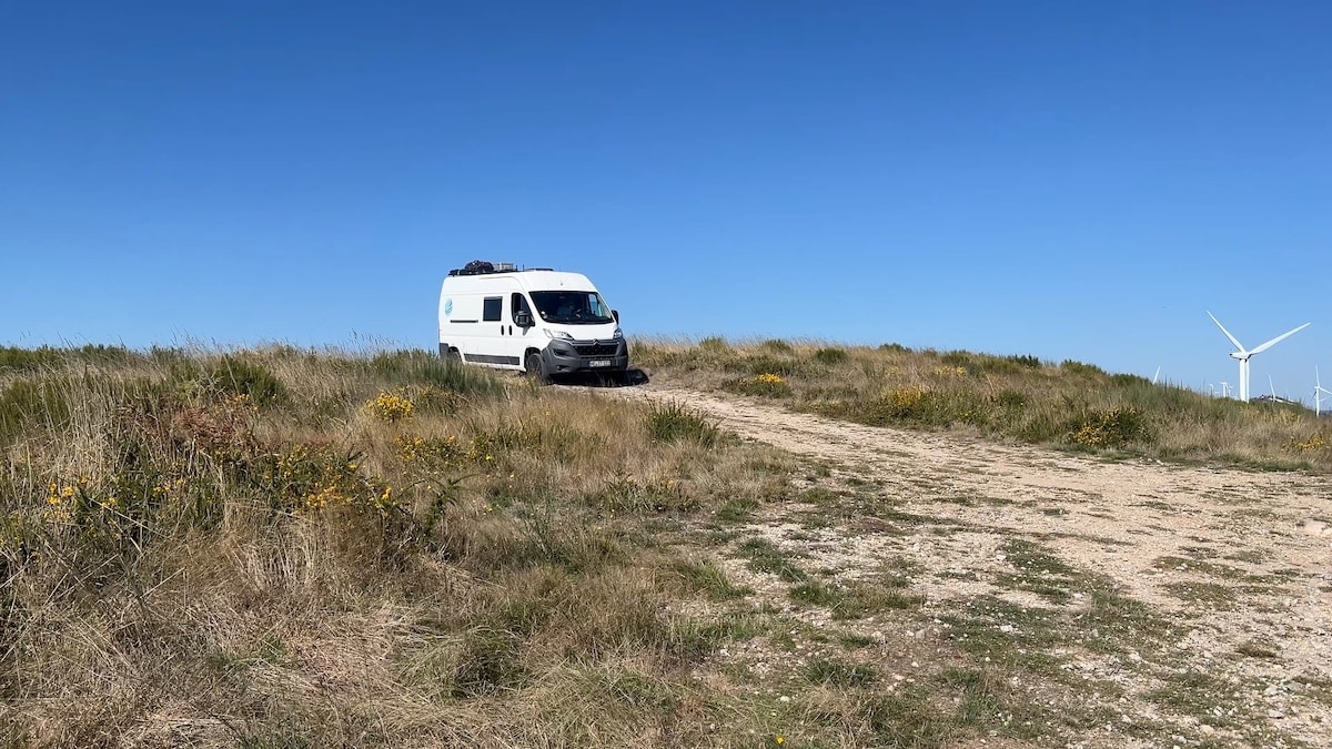 Ein weißes Wohnmobil steht auf einem grasbewachsenen, unbefestigten Weg unter strahlend blauem Himmel, im Hintergrund sind Windräder zu sehen.