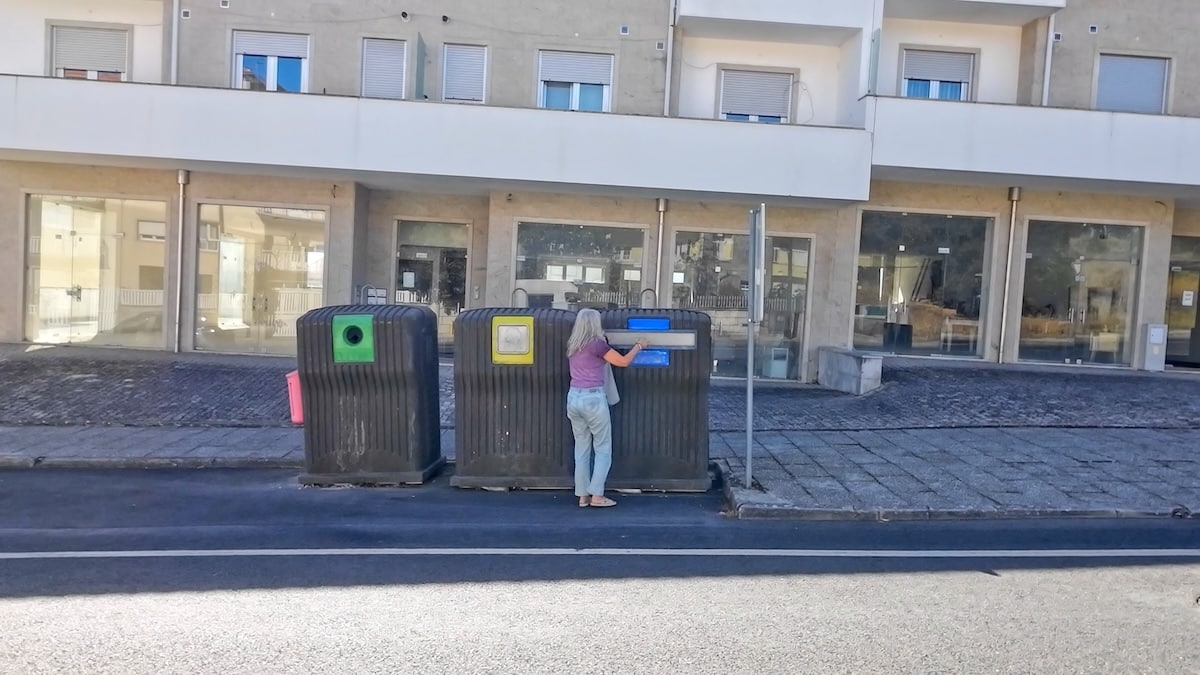 Eine Person steht auf einem Gehweg und wirft einen Gegenstand in einen Recycling-Behälter zwischen drei großen Abfallbehältern vor einem Gebäude.