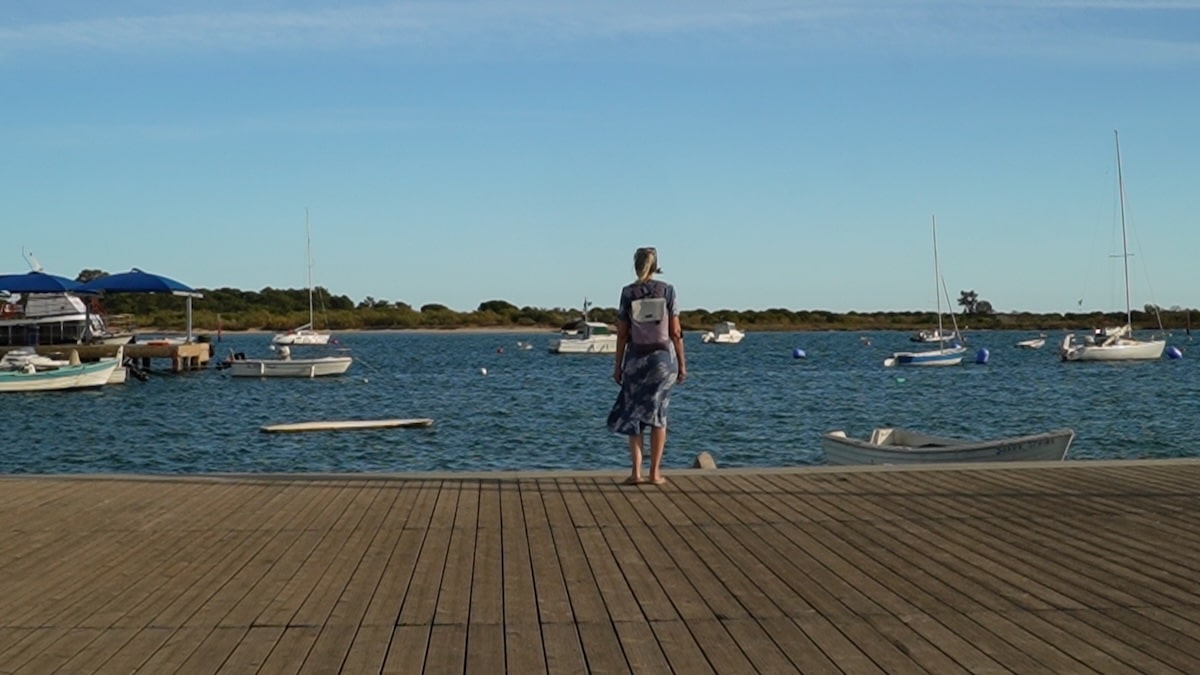 Eine Person in einem blauen Kleid steht auf einem hölzernen Steg mit Blick auf eine ruhige Bucht mit mehreren vertäuten Segelbooten unter einem klaren blauen Himmel.