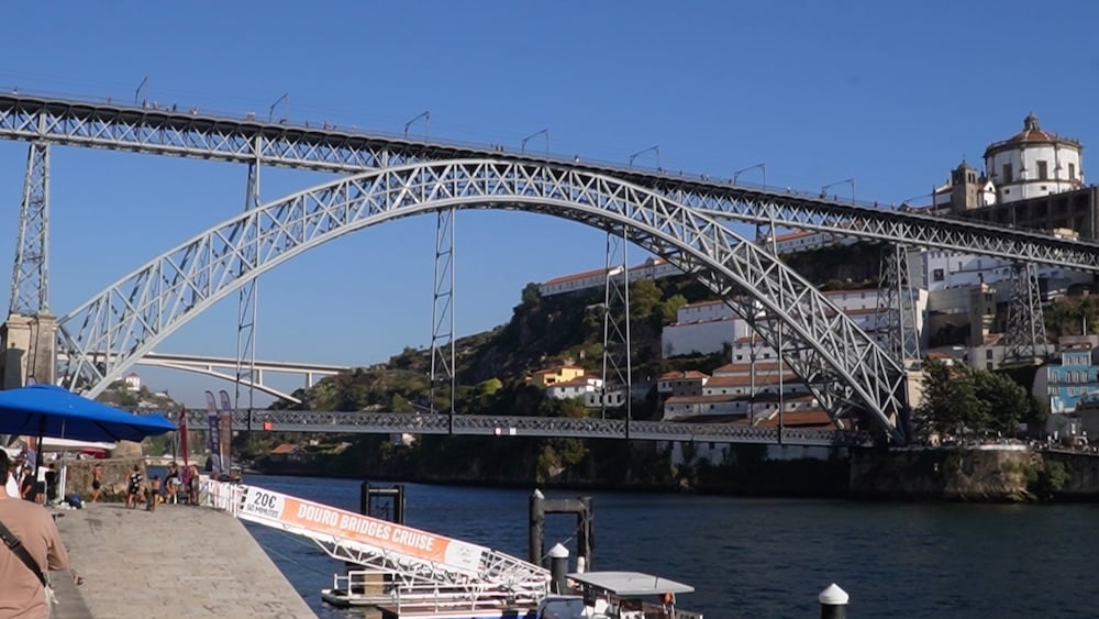 Eine große Metallbogenbrücke überspannt einen Fluss, während im Vordergrund Gebäude an einem Hang und ein angedocktes Schiff zu sehen sind, das Rundfahrten auf der Douro-Brücke anbietet.