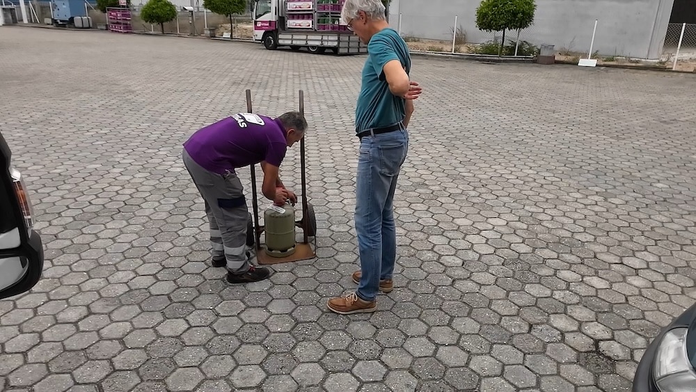 Zwei Männer stehen auf einer gepflasterten Fläche; einer bückt sich, um eine Gasflasche zu inspizieren, während der andere mit den Händen in den Hüften zuschaut.