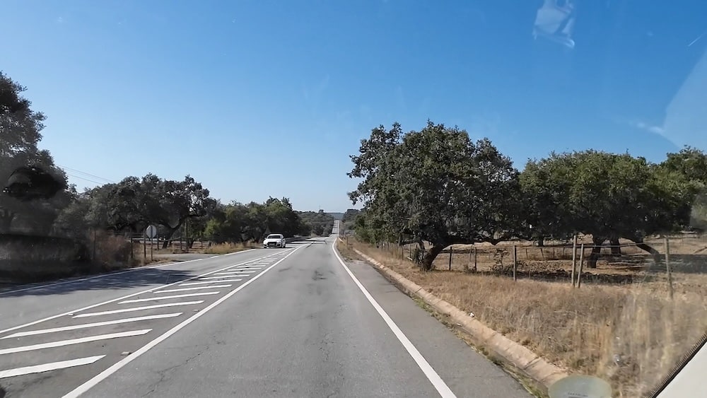 Eine zweispurige Landstraße mit markierten Linien führt durch eine trockene Landschaft mit vereinzelten Bäumen unter einem klaren blauen Himmel. Ein weißes Auto nähert sich aus der Gegenrichtung.