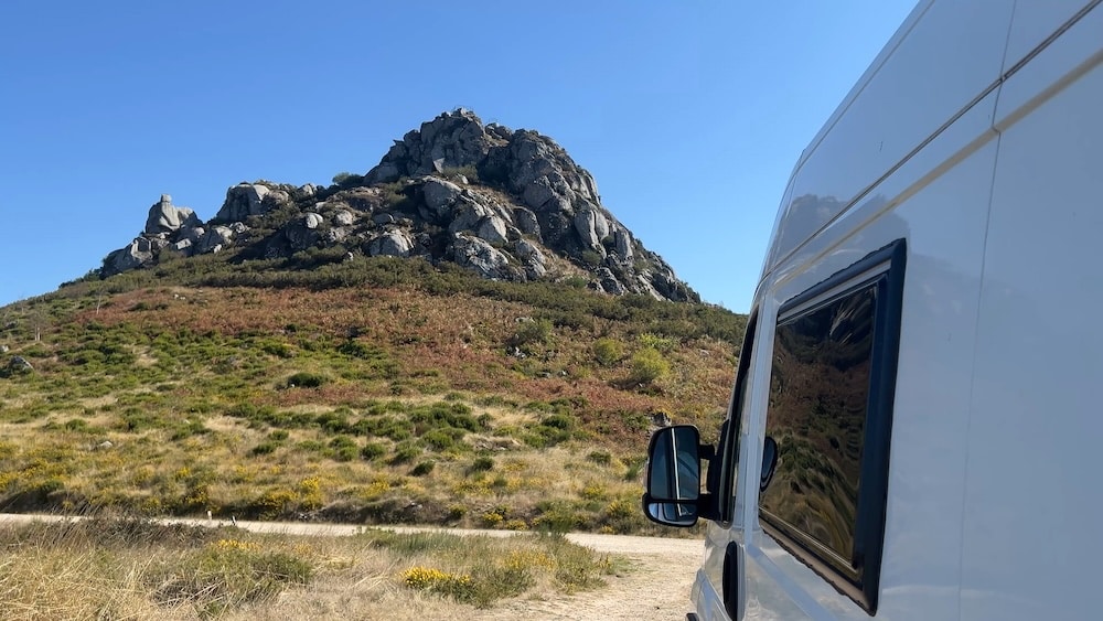 Ein weißes Wohnmobil parkt auf einer unbefestigten Straße in der Nähe eines felsigen Hügels unter einem strahlend blauen Himmel.