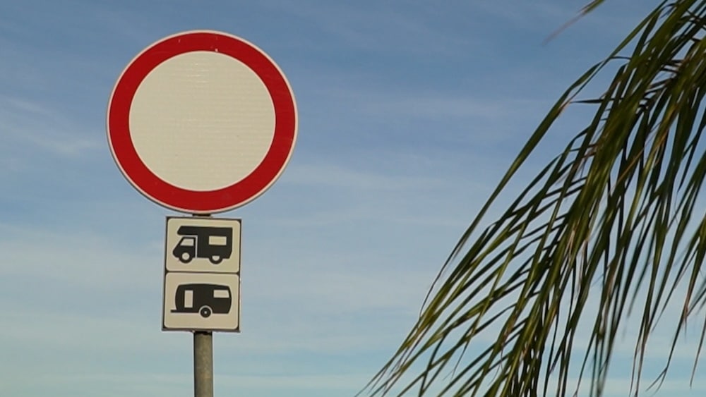 Ein Straßenschild mit einem roten Kreis über zwei Symbolen, die darauf hinweisen, dass die Einfahrt für Wohnmobile und Wohnwagen verboten ist, mit einem teilweise sichtbaren Palmwedel auf der rechten Seite.