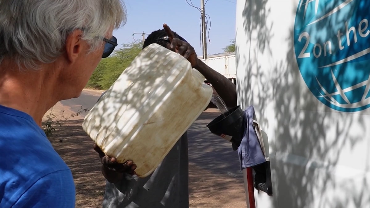 Ein Mann füllt aus einem großen, weißen Plastikkanister Wasser in den Van, während eine andere Person mit Sonnenbrille ihn beobachtet.