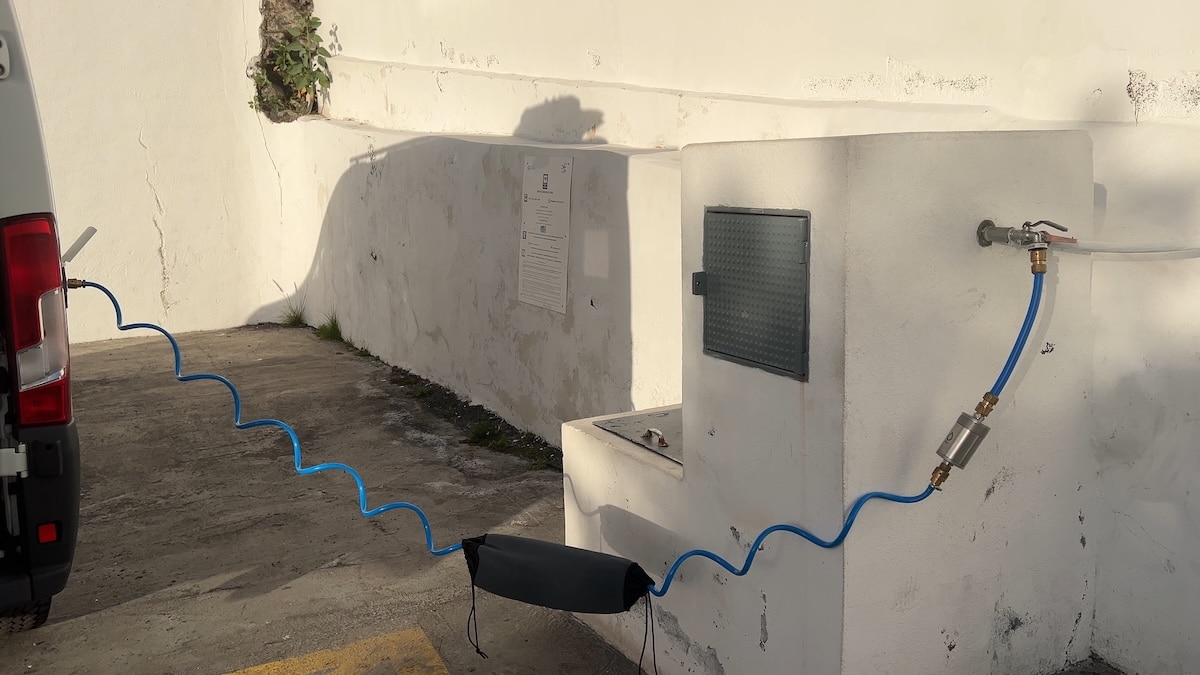 Ein blauer Schlauch verbindet einen Wasserhahn an einer weißen Wand mit einem in der Nähe geparkten Van, wobei das Sonnenlicht Schatten auf den Bürgersteig und die Wand wirft.