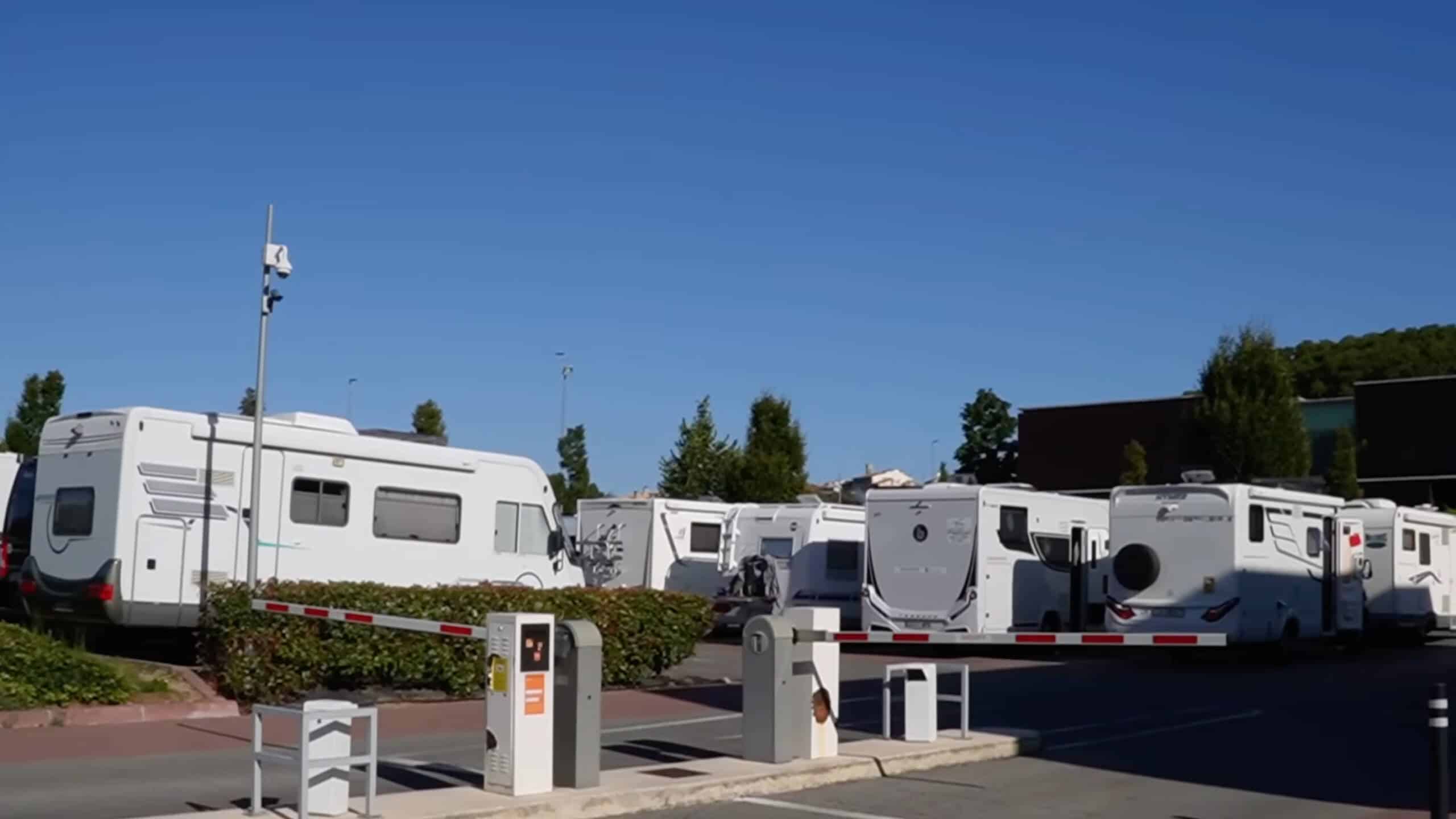 An einem klaren, sonnigen Tag stehen mehrere weiße Wohnmobile auf einem Stellplatz hinter einer Schranke. Im Hintergrund sind einige Bäume und ein Gebäude zu sehen.