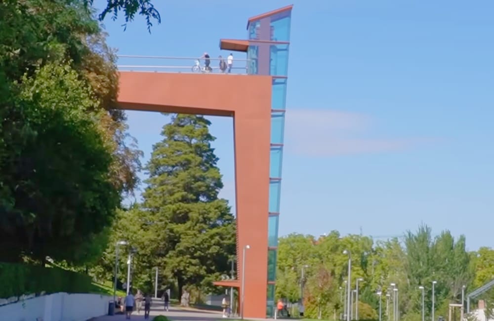 Ein hohes, modernes, orangefarbenes Gebäude mit Glasaufzug und Aussichtsplattform überragt einen Park mit Bäumen und Spazierwegen; oben und unten sind Menschen zu sehen.
