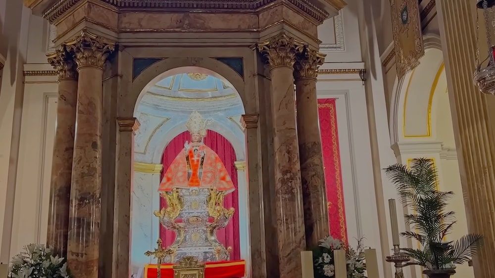 Ein reich verzierter religiöser Altar mit Marmorsäulen, einer Statue in roten und silbernen Gewändern sowie Blumenarrangements zu beiden Seiten.