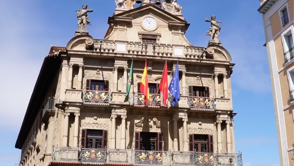 Ein prunkvolles Steingebäude mit Balkonen, einer Uhr, Statuen auf dem Dach und zahlreichen Flaggen an der Fassade vor blauem Himmel.