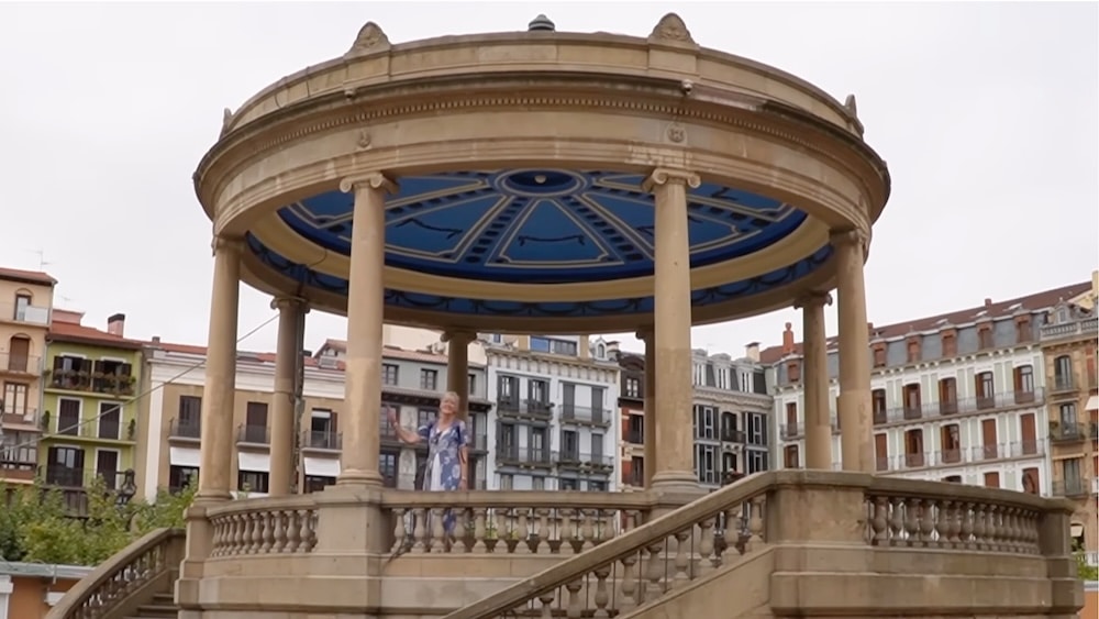 Eine Person steht in einem runden, offenen Pavillon mit blauer Decke, umgeben von Gebäuden im europäischen Stil.