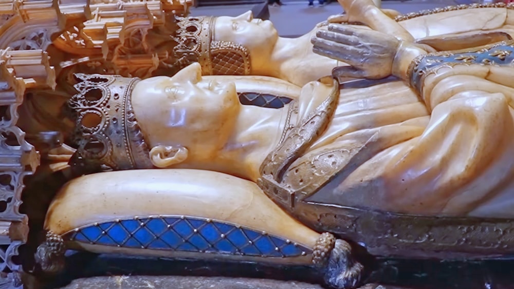 Zwei bis ins kleinste Detail ausgearbeitete Statuen von Königsfamilienmitgliedern mit Kronen liegen Seite an Seite, die Hände zum Gebet gefaltet, auf kunstvoll verzierten Kissen in einer historischen Kulisse.