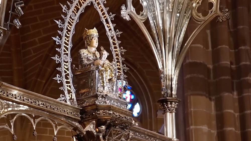 Silberstatue einer gekrönten Gestalt, die ein Kind im Arm hält und auf einem kunstvoll verzierten Sockel sitzt, in einer Kirche mit gewölbten Steinwänden und Buntglasfenstern.