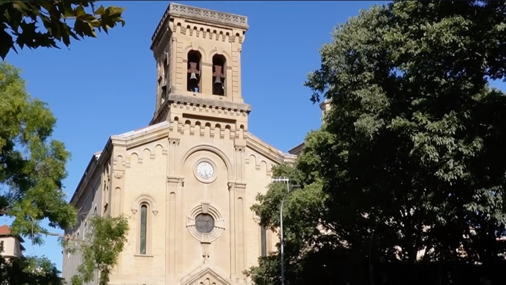 Eine beigefarbene Steinkirche mit einer Uhr und Bogenfenstern, teilweise von grünen Bäumen verdeckt, vor einem strahlend blauen Himmel.