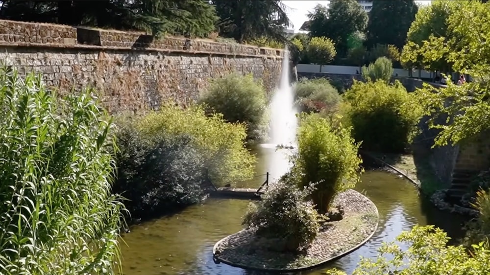 Eine Steinmauer säumt einen schmalen Bach mit einer kleinen Insel aus Sträuchern und einem Springbrunnen in der Mitte, aus dem Wasser nach oben spritzt.