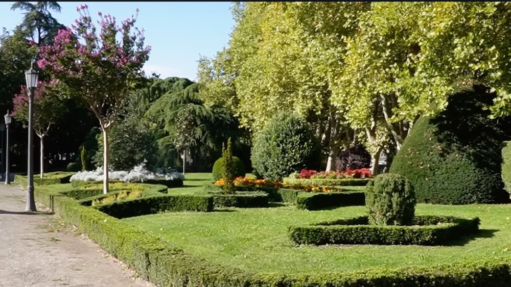 Ein gepflegter Park besticht durch geometrisch geschnittene Hecken, Blumenbeete, gestutzte Sträucher und Bäume unter strahlend blauem Himmel.