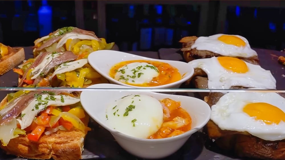 Verschiedene Pintxos auf einem Tablett, darunter Toast mit Sardellen und Paprika, Schälchen mit pochierten Eiern in Soße sowie Toast mit Spiegeleiern.