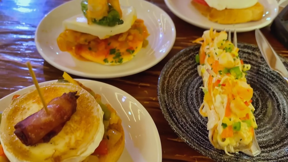 Auf einem Holztisch werden drei kleine Teller mit verschiedenen Pintxos serviert, darunter Brot mit frittierten Speisen, Ei, Gemüse und ein Spieß.
