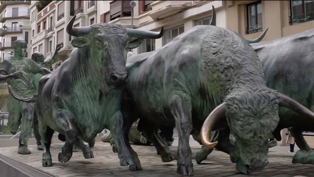 Bronzestatuen von rennenden Stieren und einem Mann sind in einer städtischen Außenkulisse ausgestellt, umgeben von Gebäuden und Menschen.