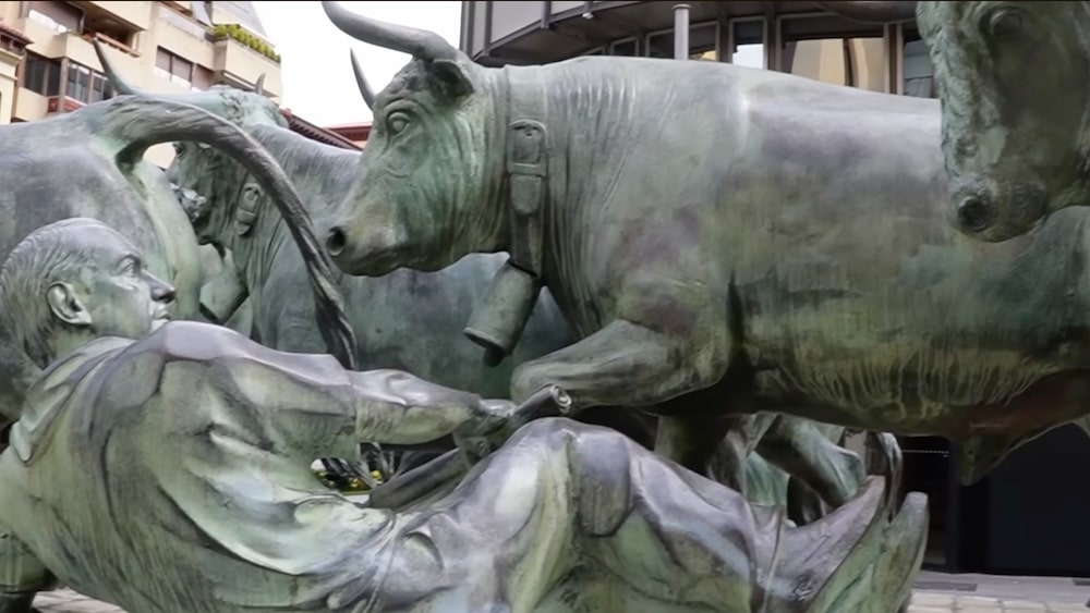 Bronzestatue eines Mannes, der auf dem Boden liegend einen Stier auf sich zurennen sieht; im Hintergrund sind weitere Stiere und Figuren in einer städtischen Umgebung zu sehen.