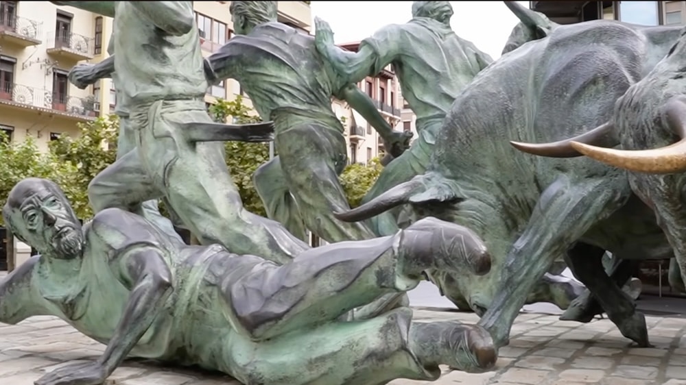 Bronzeskulptur, die Menschen zeigt, die vor Stieren fliehen, wobei ein Mann am Boden liegt; sie befindet sich auf einem städtischen Platz im Freien, umgeben von Gebäuden und Bäumen.