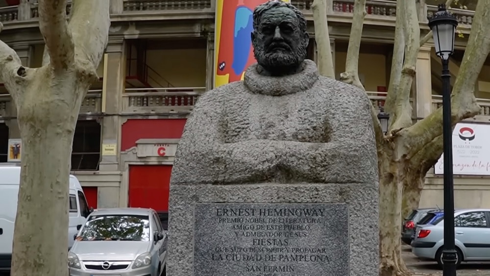 Granitstatue von Ernest Hemingway mit verschränkten Armen, die im Freien zwischen Bäumen und Gebäuden steht und auf deren Sockel eine Inschrift zu sehen ist.
