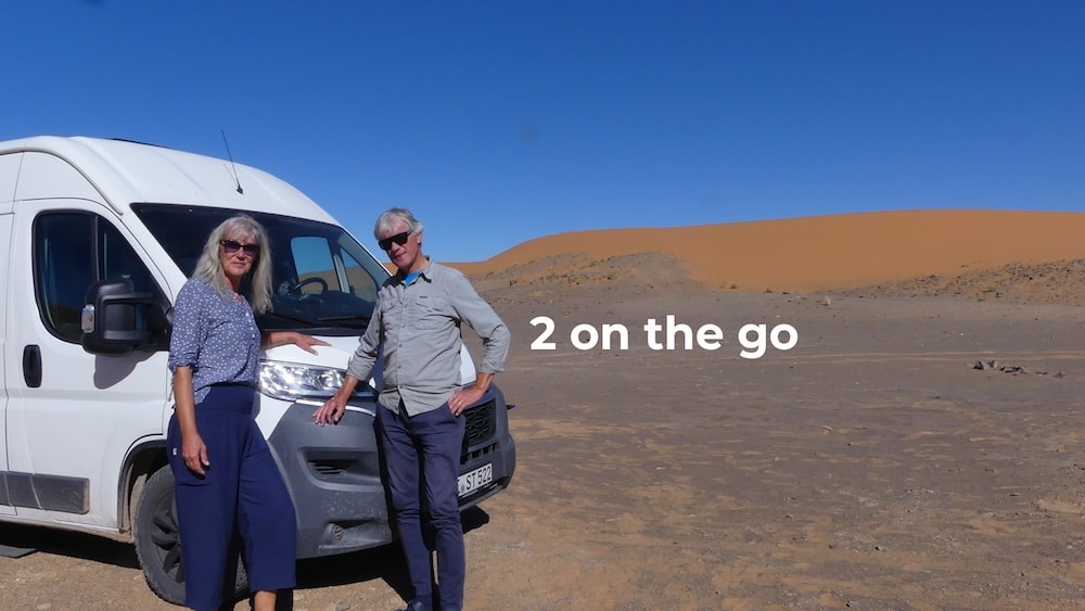 Gina und Marcus stehen neben einem weißen Van in einer Wüstenlandschaft unter strahlend blauem Himmel mit orangefarbenen Sanddünen; der Text lautet „2 on the go“.