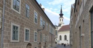 Schmale Kopfsteinpflasterstraße mit historischen Gebäuden, die zu einer Kirche mit einem hohen Kirchturm in der Ferne führt. Ein paar Leute laufen unter einem leicht bewölkten Himmel die Straße entlang, perfekt, um versteckte Budapest-Tipps zu entdecken.