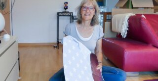 Eine Frau mit Brille und kurzen Haaren sitzt auf einem Holzboden und hält ein Stück Stoff mit einem Sternenmuster in der Hand. Sie lächelt. Im Hintergrund sind ein rotes Sofa und Kisten zu sehen.