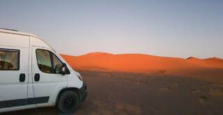 Weißes Wohnmobil steht zwischen rötlichen Dünen in der Wüste.