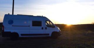 Ein weißer Van steht bei Sonnenuntergang auf einer Rasenfläche. Die Sonne steht tief am Horizont und wirft lange Schatten.